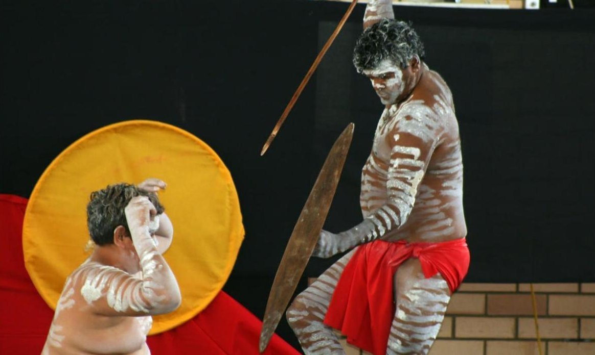A dancer with spear and shield performing in front of the Aboriginal flag