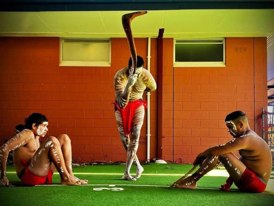 A dancer holding a boomerang aloft between two seated dancers